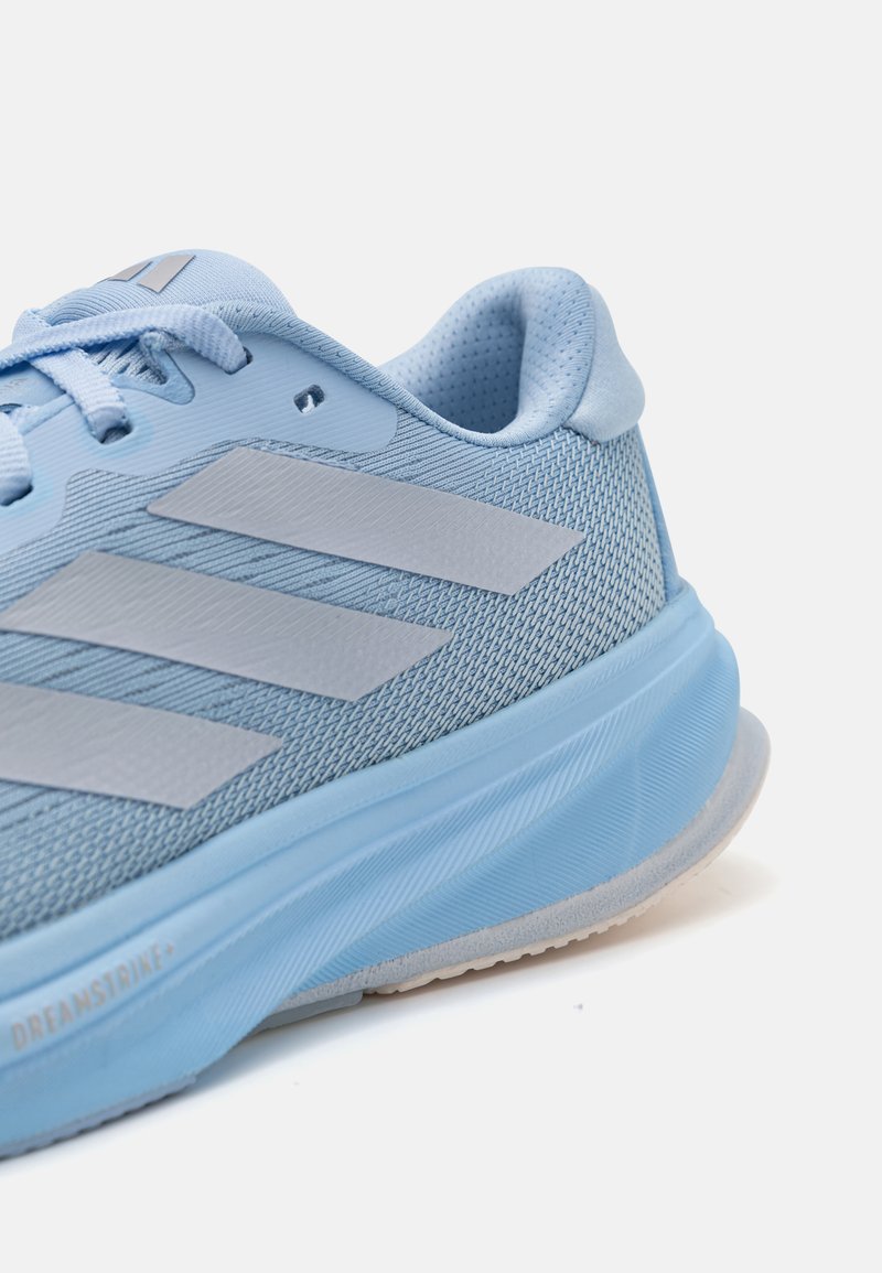 Zapatilla deportiva de color azul claro con parte superior de malla, tres franjas grises, cuello acolchado y suela de goma redondeada. Adecuada para actividades deportivas.