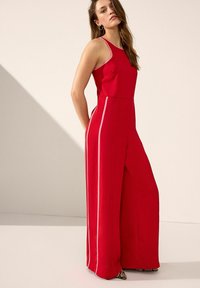 Röd byxdress med vida ben, ärmlös design och kontrasterande sidostreck. Slät tyg med figurnära overdel och avslappnade fickor.