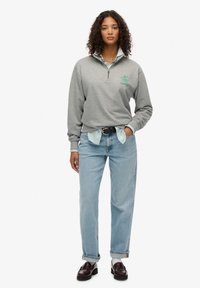 Sudadera gris con cierre de cuarto y logo verde, llevada sobre una camisa a rayas en azul claro y blanco, combinada con jeans azul claro y zapatos oscuros.