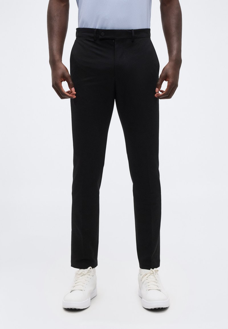 Μπροστινή όψη ατόμου που φοράει στ ενά μαύρα παντελόνια, λευκά αθλητικά παπούτσια και ανοιχτό μπλε πουκάμισο, στέκεται μπροστά σε απλό φόντο.