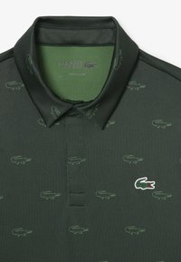 Mörkgrön polo skjorta med ton-i-ton krokodil mönster över hela plagget och en vit krokodil-logotyp. Har klassisk krage och knappslå.