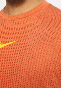 Pomarańczowa sportowa koszulka z teksturowanej tkaniny z wzorem w kratkę, okrągłym dekoltem i kontrastowym żółtym logo Nike na piersi.