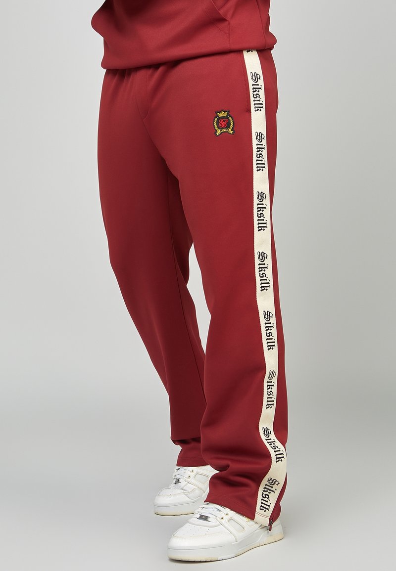 Rote Jogginghose aus weichem Stoff mit schmal zulaufender Passform. Mit einem cremefarbenen Seitenstreifen mit schwarzem Text und einem kleinen gestickten Logo am Oberschenkel.