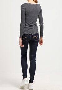 Top largo de manga larga gris con ribetes y dobladillo en forma de scallop, combinado con jeans ajustados azul oscuro y zapatillas blancas, visto desde la parte de atrás.