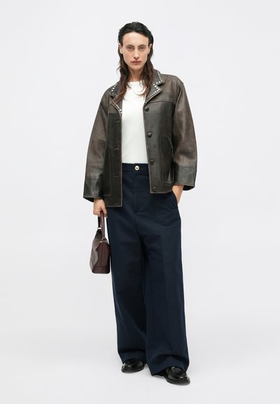 Braune Lederjacke mit Nietenkragen, weißes Hemd, dunkelblaue weite Hose, hält eine kastanienbraune Handtasche, schwarze Schuhe. Einfarbiger Hintergrund.
