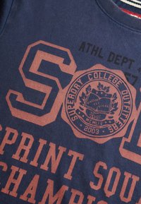 Camiseta azul marino con gran texto y gráfico en rojo-anaranjado. Presenta el sello de "SUPERDORY COLLEGE OUTFITS", fuente atlética moderna y cuello a rayas.