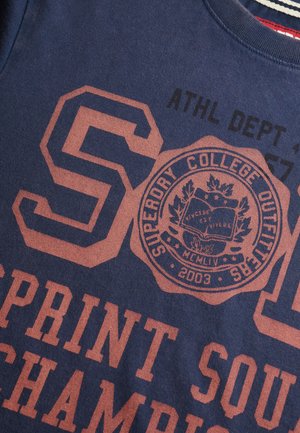 Marineblå t-shirt med stor rødlig-orange tekst og grafik. Har "SUPERDORY COLLEGE OUTFITS" emblem, moderne atletisk skrifttype og stribet halsudskæring.
