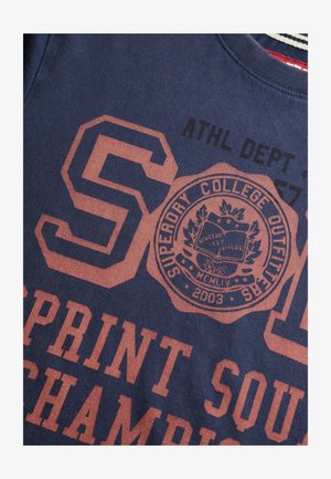 Marineblå t-shirt med stor rødlig-orange tekst og grafik. Har "SUPERDORY COLLEGE OUTFITS" emblem, moderne atletisk skrifttype og stribet halsudskæring.