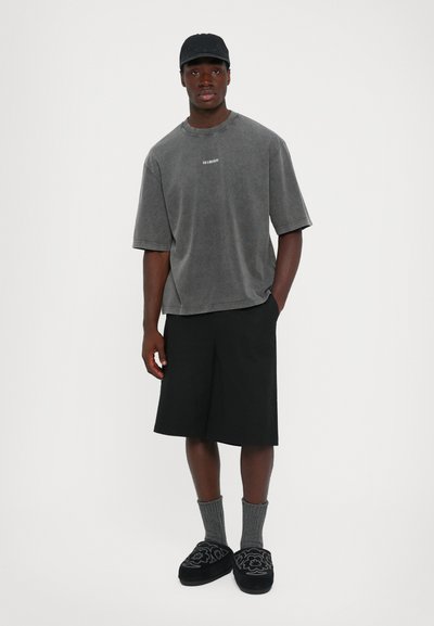 Jonge man in een gewassen grijze oversized T-shirt, zwarte shorts tot de knie, grijze sokken, zwarte geborduurde slippers en een zwarte pet.