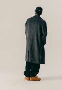 Personne portant un long manteau gris, un pantalon noir, des bottes beiges et une casquette de baseball noire, se tenant de dos contre un fond uni.