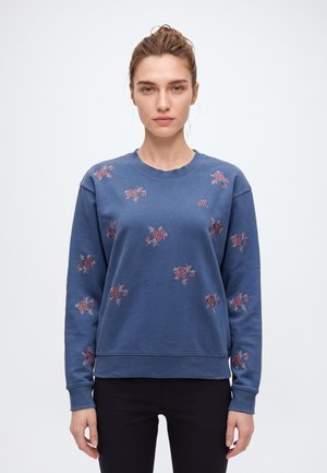 Une femme porte un sweat-shirt bleu à col rond orné de broderies florales roses, ainsi qu’un pantalon noir, sur un fond clair uni.