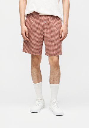 Moški, ki nosi rdeče-belo karo kratke hlače z gumbstim preklopom, bele nogavice s kratkim ovrom, bele superge in zatlačeno belo majico s kratkimi rokavi.