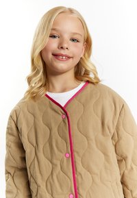 Quilted beige jakke med rosa kant og knapper; har en bølget tekstur og lukning foran. Bæres over en hvid skjorte.