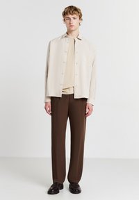 Beige Hemd mit Knopfleiste über einem beigen Oberteil, kombiniert mit braunen Hosen und dunklen Slippern. Einfaches Design, glatte Textur und neutrale Farben.