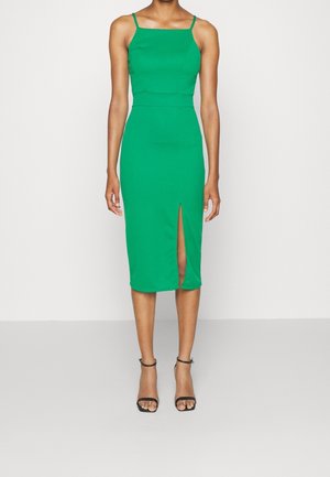 Kobieta w dopasowanej zielonej sukience midi na cienkich ramiączkach z rozcięciem z przodu, w zestawieniu z czarnymi szpilkami z paskiem wokół kostki.