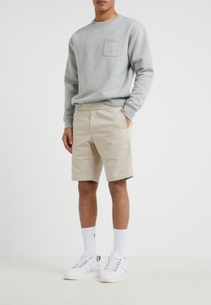 Uomo che indossa una camicia a maniche lunghe grigio chiaro con taschino sul petto, pantaloncini beige, calzini bianchi fino alla caviglia e scarpe da ginnastica bianche, in piedi davanti a uno sfondo semplice.