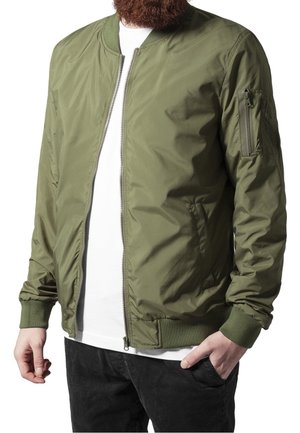 Homme portant une veste bomber vert olive sur une chemise blanche et un pantalon noir, une main dans la poche et l'autre détendue.