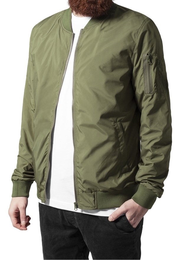 Homme portant une veste bomber vert olive sur une chemise blanche et un pantalon noir, une main dans la poche et l'autre détendue.