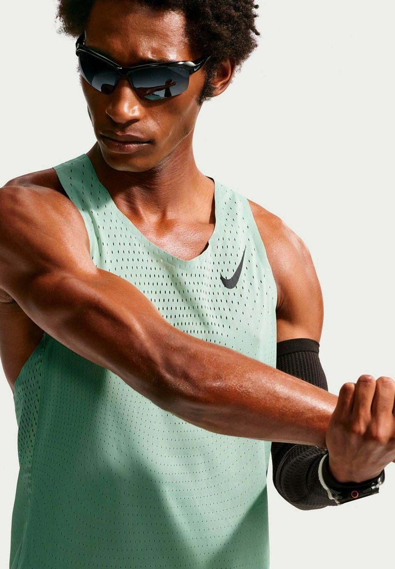 Athlète portant un débardeur Nike vert clair et une manche noire s'étire le bras, portant des lunettes de soleil et un tracker de fitness.