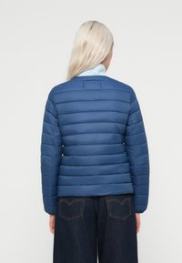 Modrá puffer bunda s vertikálnou prešívanou textúrou, okrúhlym golierom a skráteným strihom, v kombinácii s tmavými denimovými širokými nohavicami.