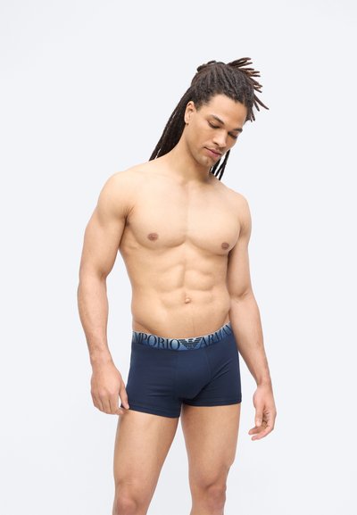 Μυώδης άνδρας με dreadlocks που φοράει μπλε ναυτικό boxer briefs Emporio Armani, στέκεται μπροστά σε απλό φωτεινό φόντο, κοιτώντας προς τα κάτω.