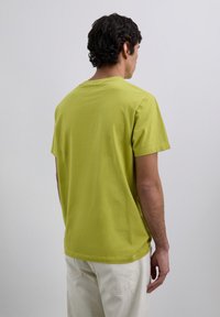 Homme avec des cheveux bouclés foncés portant une chemise à manches courtes vert lime et un pantalon beige clair, tourné le dos contre un fond uni.