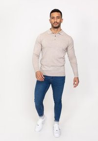 Beige pitkähihaisen poolopaidan, jossa on napillinen kaulus, yhdistetty sinisiin slim fit -farkkuihin ja valkoisiin lenkkareihin. Tasainen tekstuuri, rento malli.