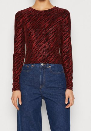 Femme portant un haut ajusté à manches longues rouge et noir à motifs, associé à un jean bleu taille haute sur fond blanc.