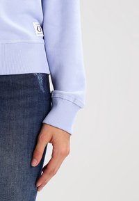 Felpa lilla con polsini e orlo a costine, texture morbida, etichetta di lato, indossata sopra un denim scuro. Mano appoggiata sul tessuto.