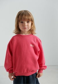 Jasnoróżowa bluza z prążkowanymi mankietami i ściągaczem u dołu, z małym białym logo. Luźny krój z długimi, lekko bufkami rękawami.