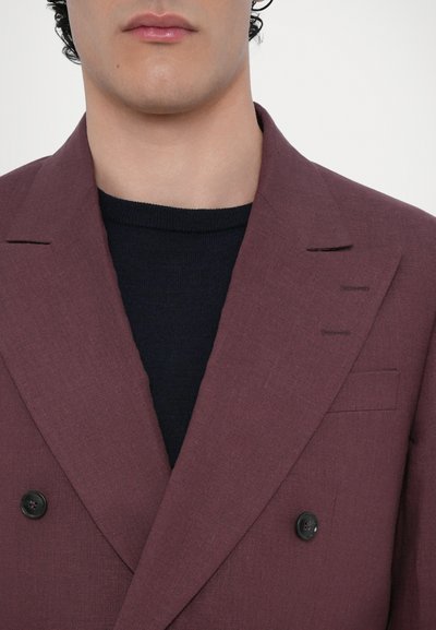 Doppelt genähter Blazer aus kastanienbraunem Leinen mit schwarzen Knöpfen, Reverskragen und strukturiertem Stoff. Getragen über einem dunkelblauen Strickhemd.