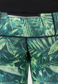 Leggings con un patrón de hojas tropicales en verde y negro. Hechos de tela elástica con costuras negras y un bolsillo en la cinturilla.