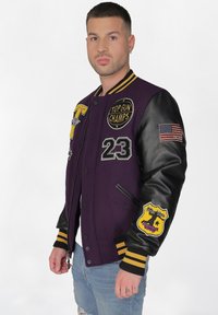 Violetin harjoittelutakki, jossa on mustat nahkahihat, ja siinä on useita tarroja, mukaan lukien "TOP GUN CHAMPS" ja Yhdysvaltojen lippu. Resorihihansuut, joissa on keltaiset raidat.