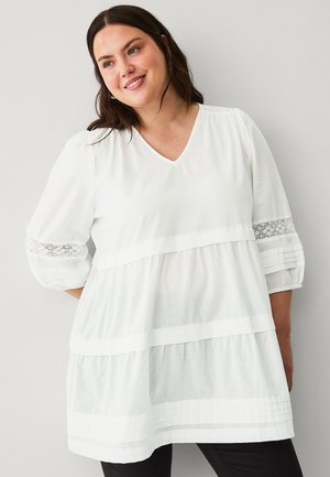 Uśmiechnięta kobieta o ciemnych włosach, ubrana w białą, warstwową bluzkę z koronkowymi detalami na bufiastych rękawach na jasnym, jednolitym tle.