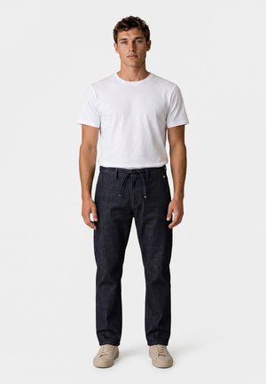 Jonge man die recht vooruit staat, draagt een effen wit t-shirt, donkerblauwe broek met trekkoord en beige sneakers op een witte achtergrond.