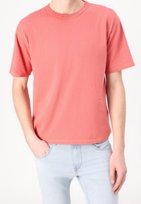 T-shirt in cotone colore corallo con scollo rotondo, maniche corte, vestibilità comoda, con un'etichetta logo sottile e dettagli di cuciture a contrasto. Abbinata a jeans azzurri.