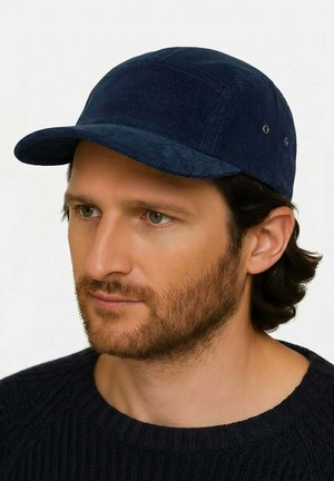 Man with short beard and medium-length dark hair wearing a dark blue corduroy baseball cap and black knitted sweater, looking slightly left.