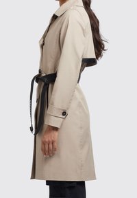Beige trenchcoat med bältad midja, med knapplist framtill, långa ärmar och en sidoklyvning. Svarta detaljer på bältet.