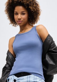 Νέα γυναίκα με σγουρά μαλλιά, φοράει μπλε αμάνικο τοπ, μαύρο δερμάτινο μπουφάν περασμένο στους ώμους και ανοιχτό μπλε πουκάμισο δεμένο γύρω από τη μέση.