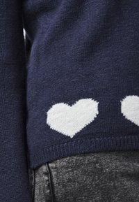 Pull en tricot bleu marine avec trois motifs de cœurs blancs à l'ourlet, en tissu texturé. Associé à un pantalon gris à motifs.