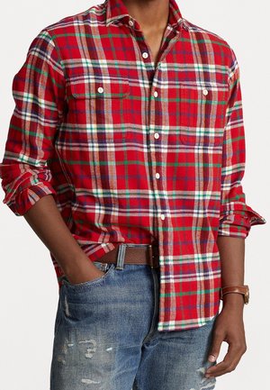 Homme portant une chemise à carreaux rouge, verte et blanche avec les manches retroussées, un jean bleu usé, une ceinture marron et une montre-bracelet avec un bracelet en cuir marron.