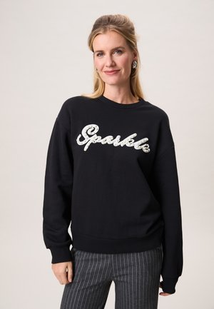 Vrouw met blond haar draagt een zwarte trui met het woord 'Sparkle' in witte parels en grijze pantalon met krijtstreep, glimlachend tegen een effen achtergrond.