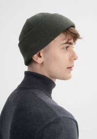 Gebreide beanie in donkergroen, geribbeld textuur, gedragen met een hooggesloten grijze trui, met een zijaanzicht tegen een witte achtergrond.