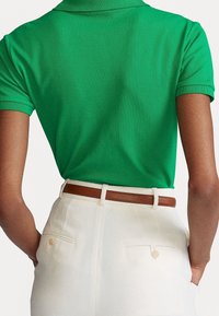 Grön piké med ribbad krage, kombinerad med hög midja i krämfärgade byxor och ett brunt läderbälte, som visar en mjuk textur och skräddarsydd passform.