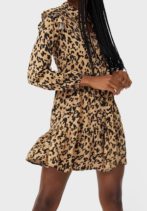 Femme portant une robe beige à manches longues avec des taches noires et des détails à volants, debout les mains jointes devant elle.