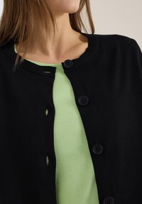 Gros plan d'une personne portant un haut vert clair sous un cardigan noir boutonné avec quatre boutons visibles.