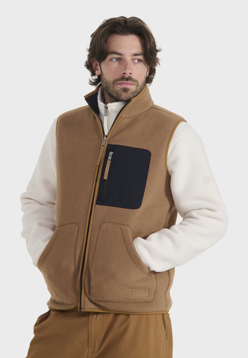 Chaleco de forro polar en marrón, con un bolsillo negro con cremallera, dos bolsillos frontales y un cuello alzado. El interior es de color crema.