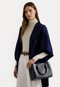 Žena oblečená v béžovém roláku, širokých bílých kalhotách s hnědým opaskem, námořnickém otevřeném cardiganě, držící černou koženou kabelku se zlatými akcenty.