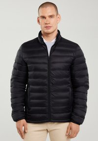 Joven con chaqueta negra acolchada con cremallera frontal y pantalones beige, posando frente a un fondo liso y claro.