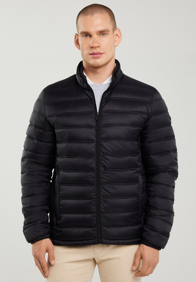 Joven con chaqueta negra acolchada con cremallera frontal y pantalones beige, posando frente a un fondo liso y claro.